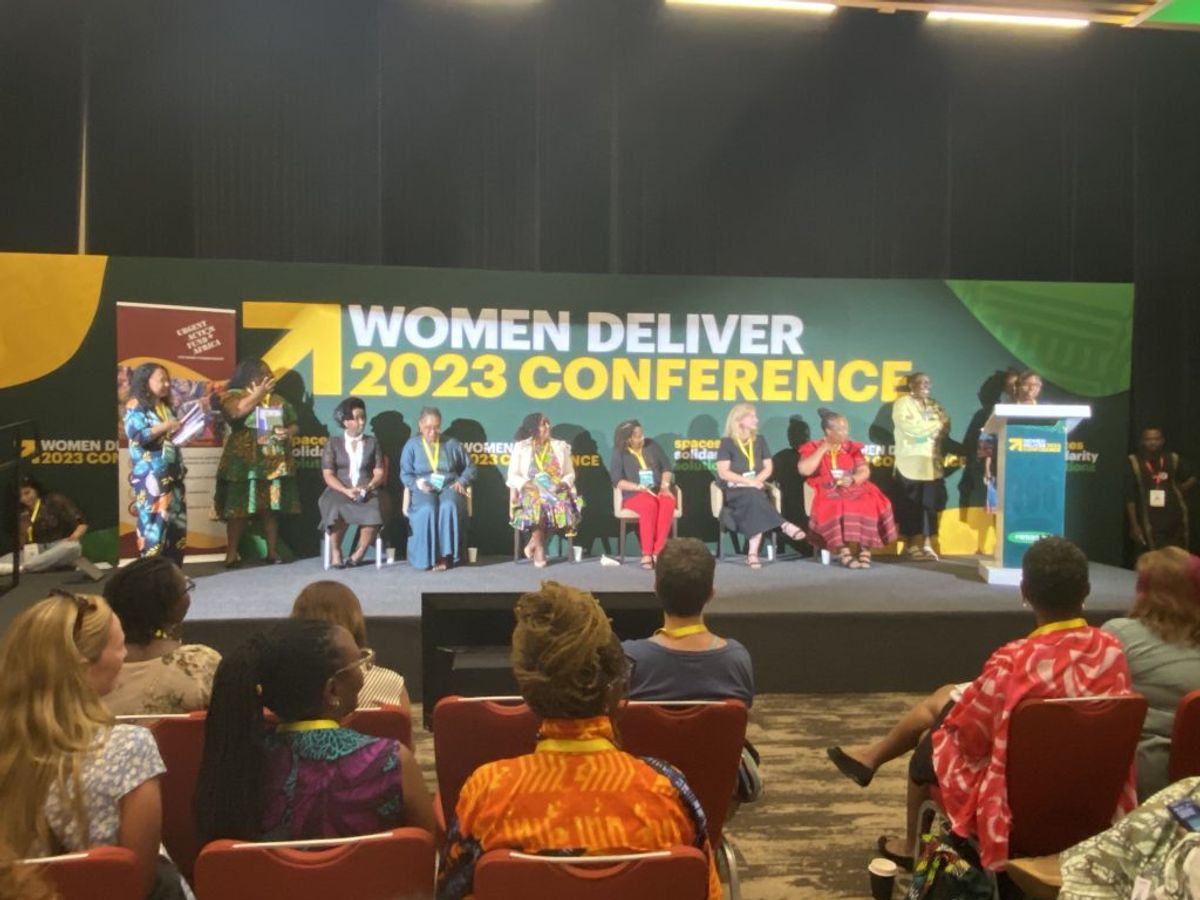 A group of people sit and stand on stage. One is behind a podium. Women Deliver banner in background